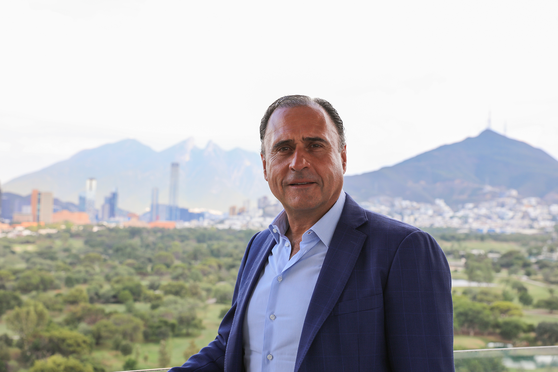 Un hombre con un traje azul se encuentra en un balcón con vistas a un paisaje urbano con montañas al fondo.