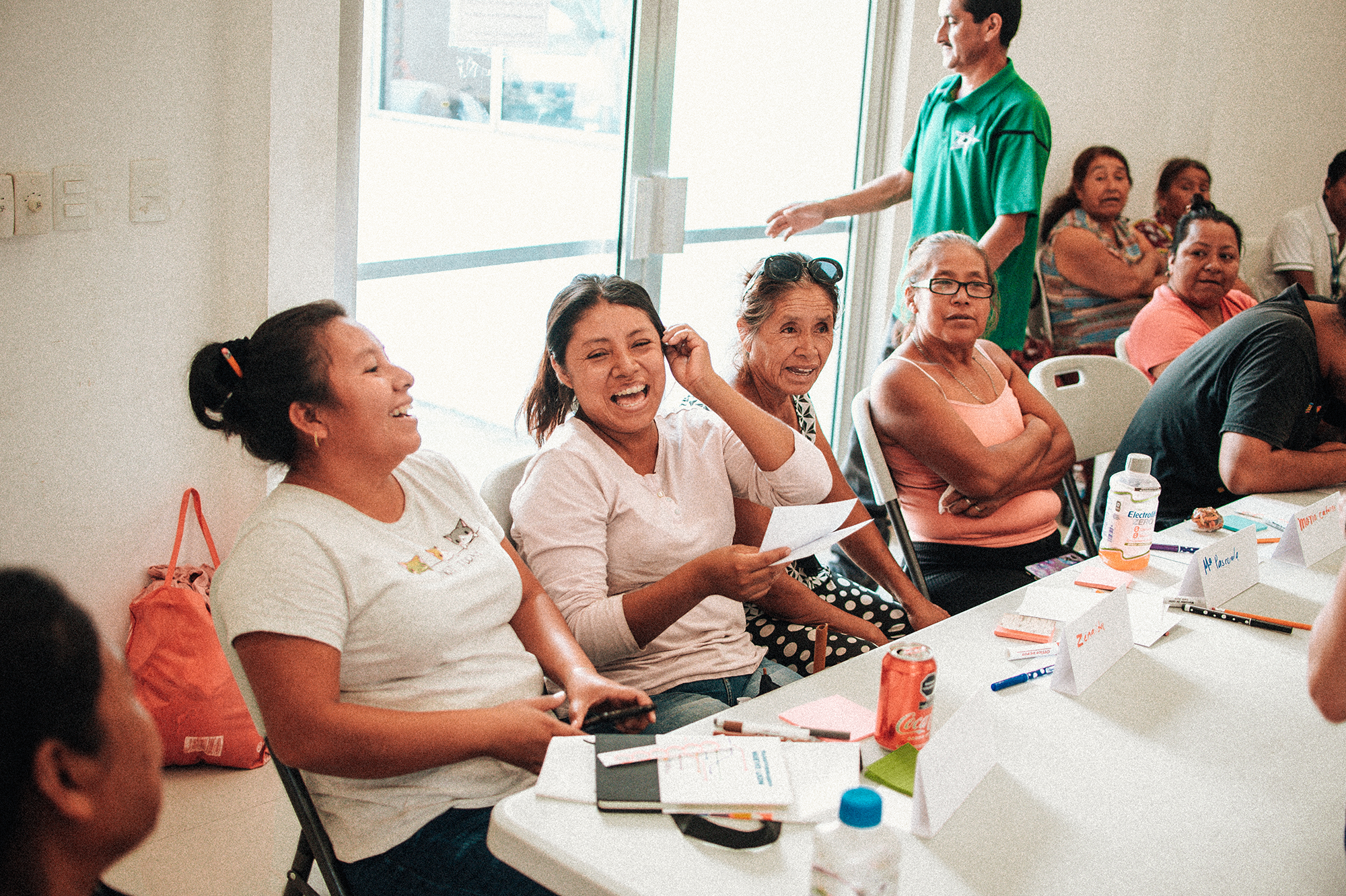 Un grupo de mujeres se sientan alrededor de una mesa, sonriendo y riendo durante una reunión. Sobre la mesa hay papeles, bebidas y tarjetas con nombres. El ambiente es informal y amistoso, con algunas personas más al fondo.