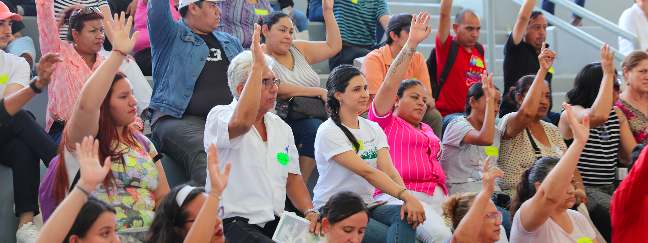 Un grupo de personas participando activamente en una asamblea, levantando la mano para expresar su opinión o voto, reflejando organización comunitaria, diálogo y compromiso colectivo.